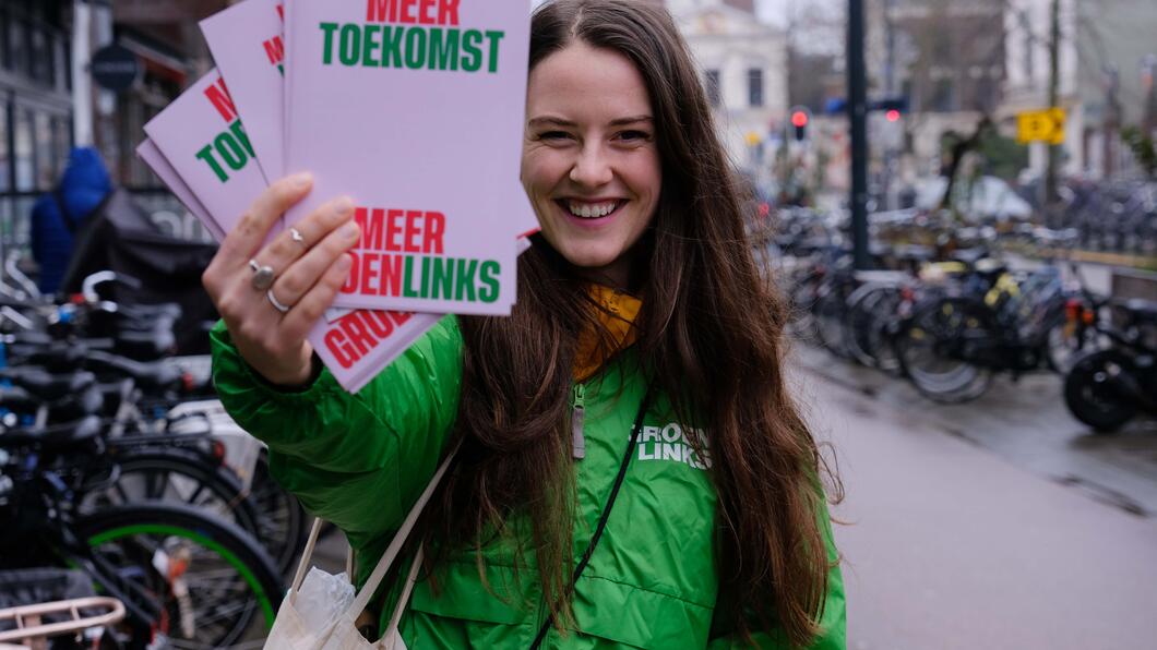 Witte vrouw met lang bruin haar heeft een groenlinks fel groen jack aan. Ze houdt groenlinks flyers aan de camera toe en glimlacht breed. Ze staat op een straat in een stad met fietsen en gebouwen op de achtergrond.