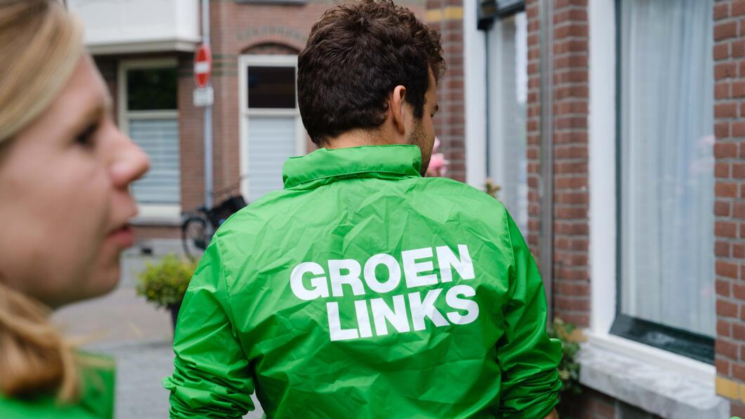 Witte man met kort bruin haar van de rug af gezien met groen groenlink jasje aan op straat naast een huis. Blonde witte vrouw onscherp op voorgrond van zijprofiel gezicht.